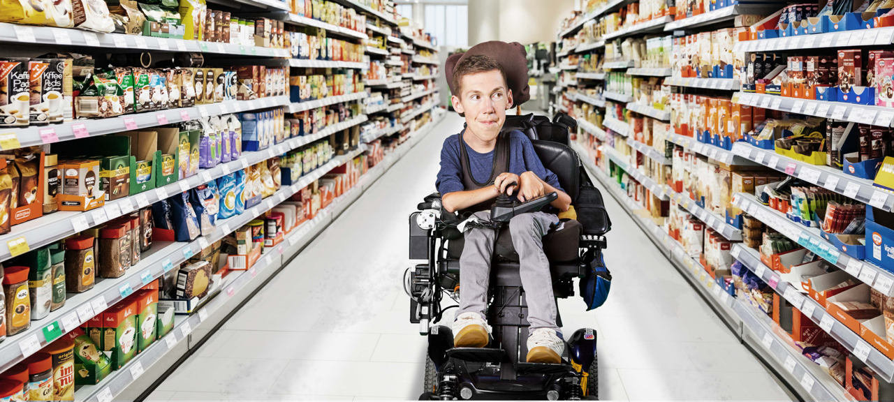 Shane est assis dans son fauteuil roulant, au milieu d’une allée de supermarché remplie de marchandises.