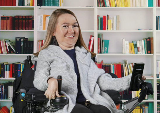 Brianna poses in her wheelchair in front of a bookshelf filled with hundreds of books. She is smiling directly at the camera.