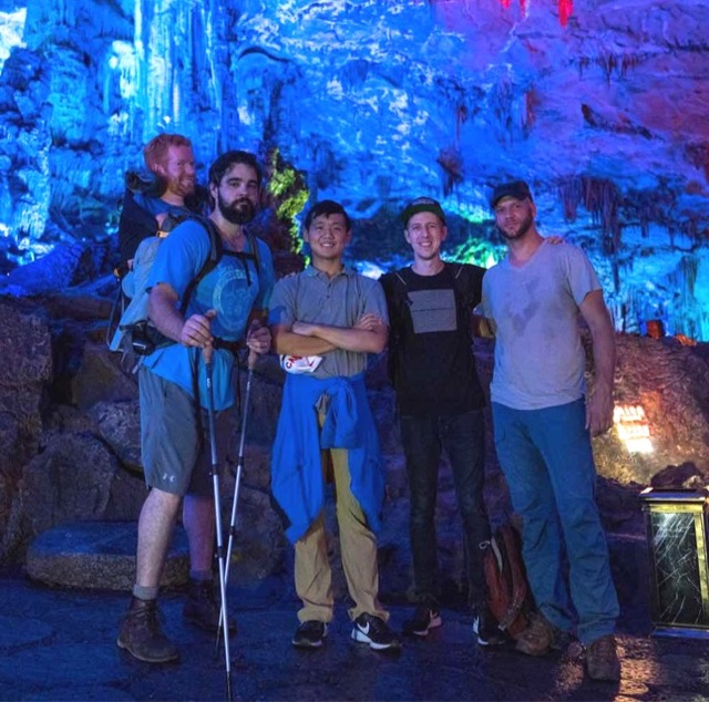 Kevan explores the Reed Flute Caves in Guilin, China with four of his friends. Kevan is being carried in his backpack by one of his companions as they all stand beside each other looking straight into the camera.
