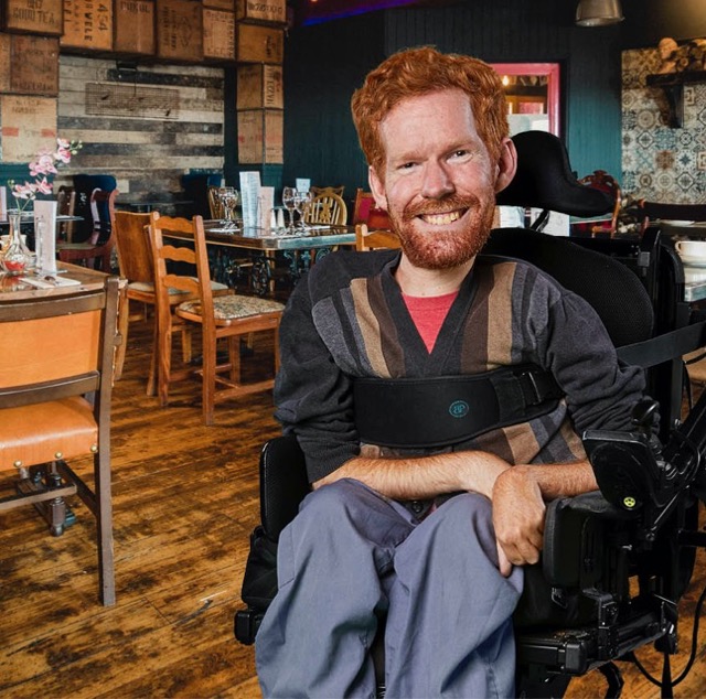 Kevan sits in his wheelchair at his favourite local coffee shop. The image shows him smiling in the middle of the shop with wooden furniture, table flowers and warm lighting.