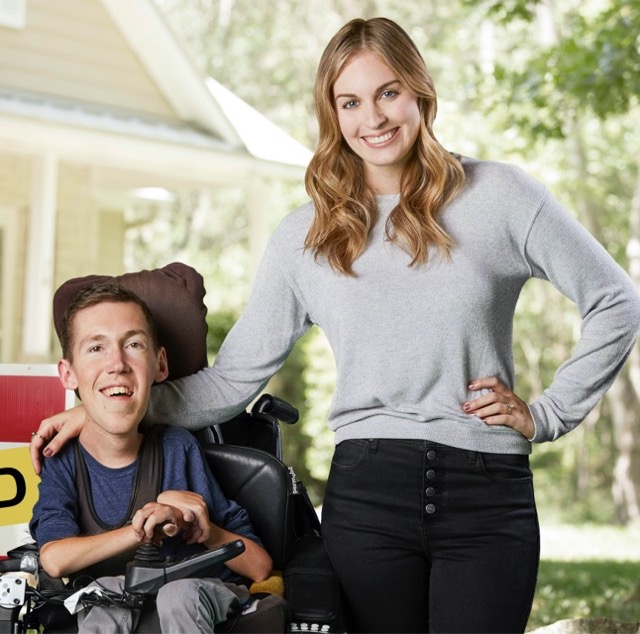 Shane and Hannah smile in front of their newly purchased home with a sold sign next to them. Shane sits in his wheelchair and Hannah stands beside him with her hand on his shoulder.